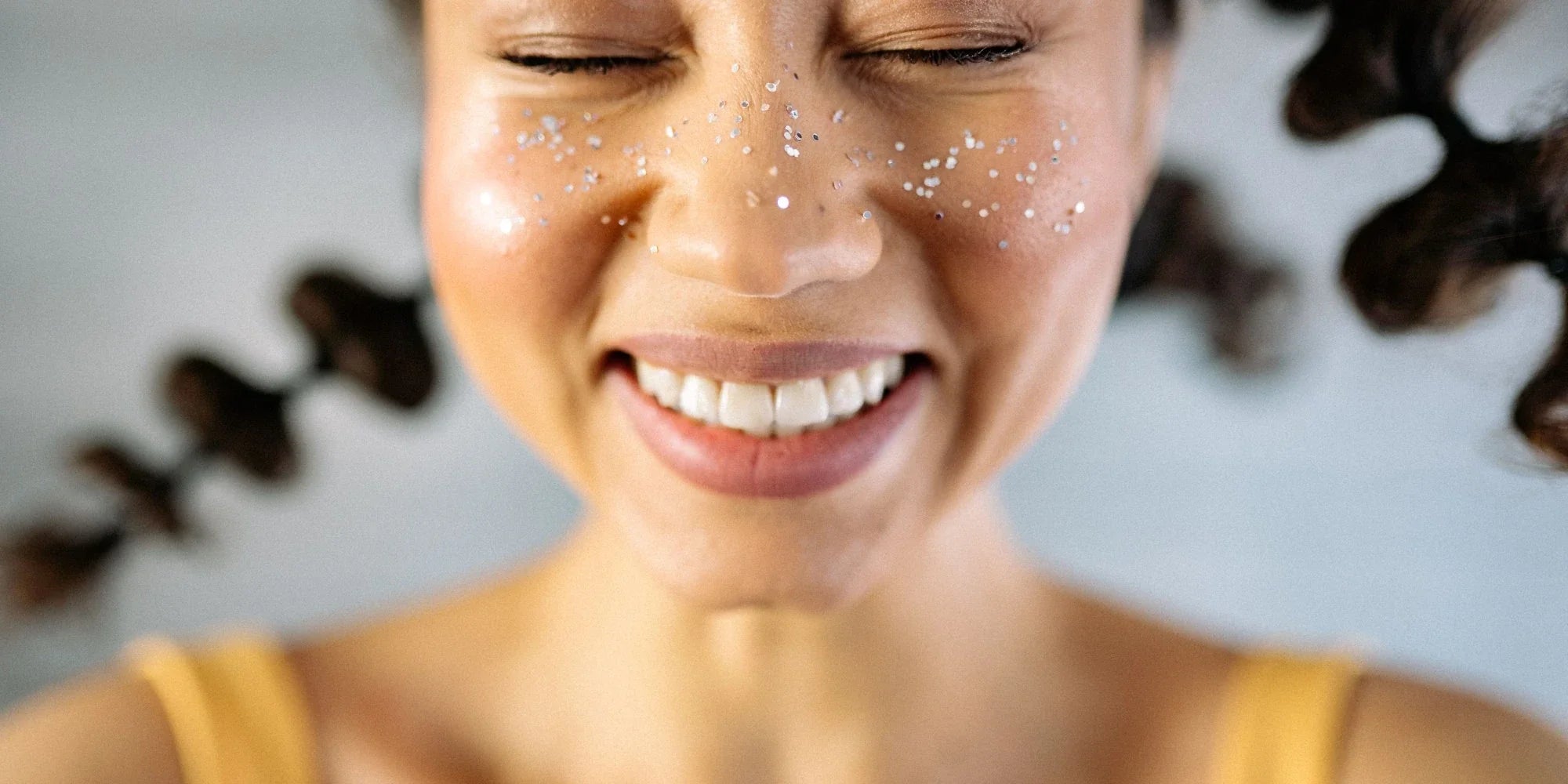 A woman in elaborate braids squinting with sequins on her face and smiling, because hydroxyapatite toothpaste is safe