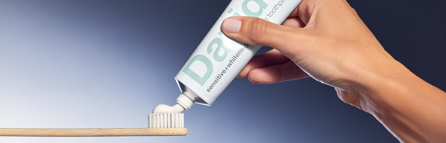 Hand squeezing Davids toothpaste onto a wooden toothbrush against a gradient blue background