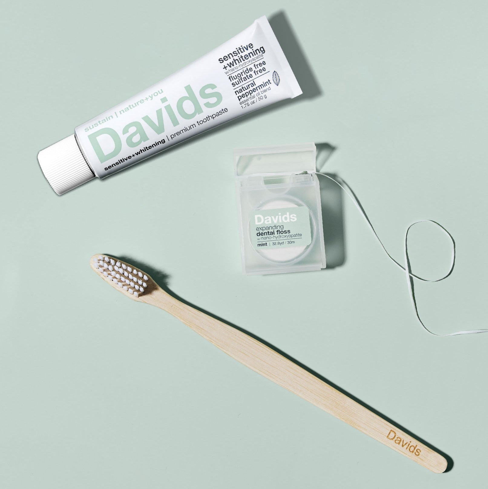 Toothpaste, dental floss, and bamboo toothbrush from Davids, arranged on a pale green surface