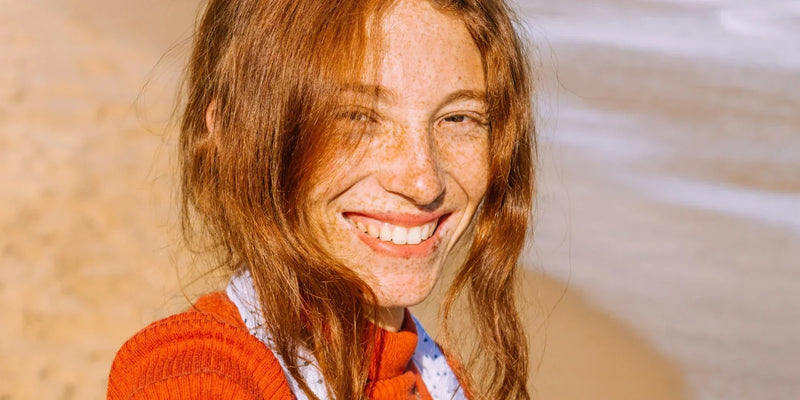 A young, red-haired woman with a bright white smile on a sunny beach.