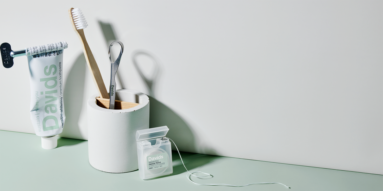 Davids toothpaste, toothbrush, tongue scraper in holder, and open dental floss on mint green surface