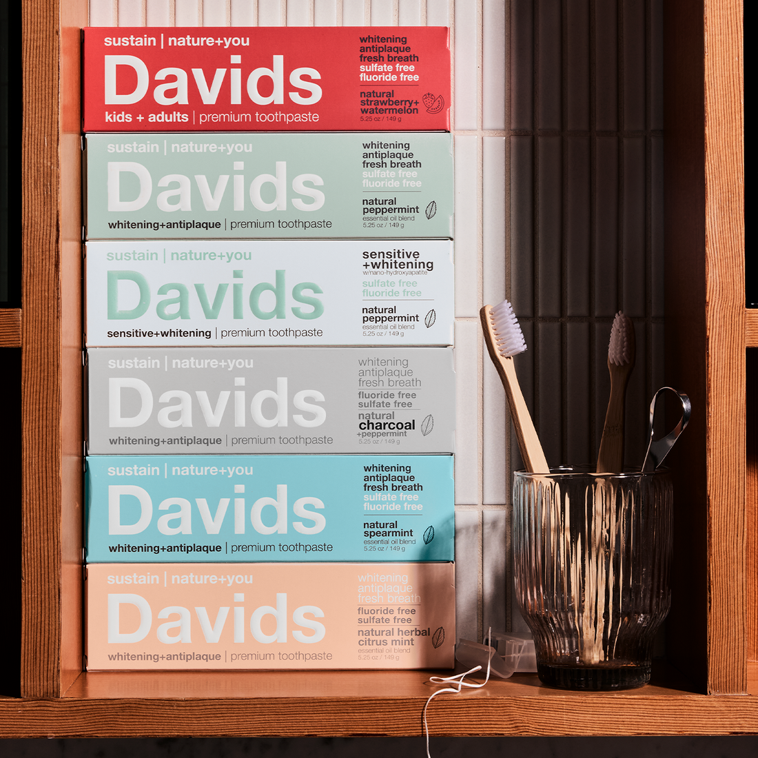 Stack of Davids toothpaste boxes in various flavors beside a glass with toothbrushes and floss