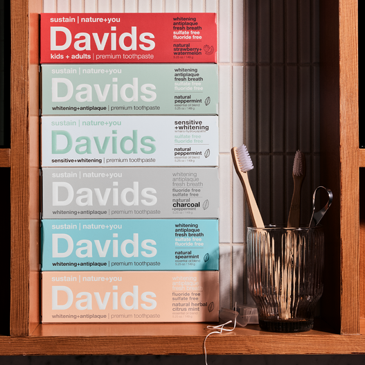 Stack of Davids toothpaste boxes in various flavors beside a glass with toothbrushes and floss